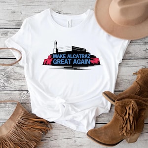 May include: White t-shirt with a graphic of Alcatraz Island and the text "Make Alcatraz Great Again". The shirt is laid out on a wooden surface with a brown hat and brown boots.