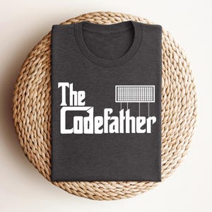 May include: Dark gray t-shirt with the text "The Codefather" in white, featuring a stylized graphic. The shirt is folded on a woven, circular placemat.