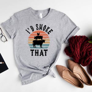 May include: A light gray t-shirt with the text "I'd Smoke That" and a graphic of a chicken, pig, and cow stacked on top of each other. The design is set against a retro sunset background. The shirt is styled with accessories.