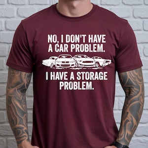 May include: Maroon t-shirt with the text "NO, I DON'T HAVE A CAR PROBLEM. I HAVE A STORAGE PROBLEM." in white. The shirt features a graphic of four cars in white. A classic car, a modern sports car, and two other car models are visible.