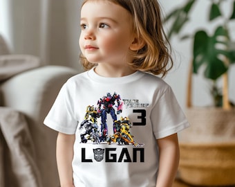 May include: White t-shirt with a Transformers graphic featuring Optimus Prime and other robots. The shirt also has the text "Transformers" and the number "3" in black. The name "Logan" is printed in black below the graphic.
