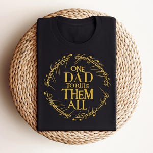 May include: Black t-shirt with a gold design that reads "One Dad to Rule Them All" in a font reminiscent of the Lord of the Rings.
