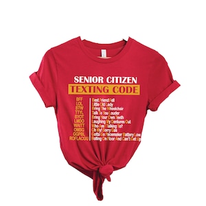 May include: Red t-shirt with the text "SENIOR CITIZEN TEXTING CODE" in white and yellow. The shirt features a list of texting acronyms and their meanings, such as "BFF" for "Best Friend Fell".