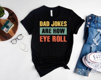 May include: Black t-shirt with the text "DAD JOKES ARE HOW EYE ROLL" in yellow, green, and orange. The shirt is paired with blue jeans and white sneakers. Other accessories include glasses and a camera.