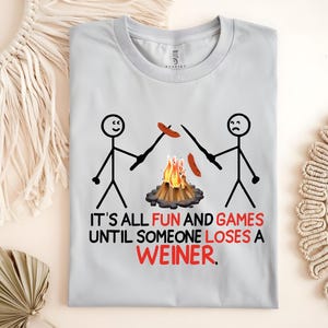 May include: A gray t-shirt with a stick figure illustration of two people roasting hot dogs over a campfire. The text reads "It's all fun and games until someone loses a weiner."