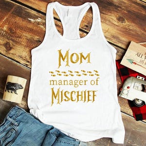 May include: White racerback tank top with gold text that reads "Mom manager of Mischief". The shirt is displayed on a wooden surface with a pair of blue jeans, a wooden block with a bear illustration, and other decorative items.