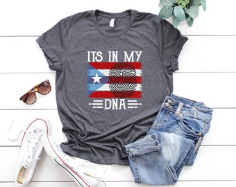 May include: A dark gray t-shirt with the text "ITS IN MY DNA" and a graphic of the Puerto Rican flag with a fingerprint heart design. The shirt is paired with blue jeans and white sneakers.