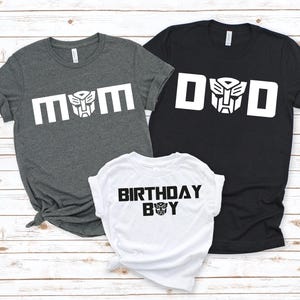 May include: Three gray, black, and white t-shirts with a Transformers theme. The shirts say "Mom", "Dad", and "Birthday Boy" with a robot head graphic.