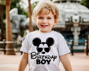 May include: A white t-shirt featuring a black graphic of Mickey Mouse wearing sunglasses, with the words "Birthday Boy" in a stylized font. The shirt is worn by a young child with blond hair, smiling at the camera.