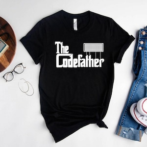 May include: Black t-shirt with the white text "The Codefather" and a graphic of a grid. The shirt is a crew neck and appears to be made of a soft material. The shirt is displayed on a light background.