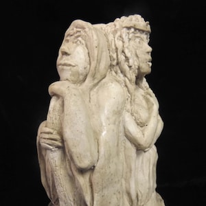 May include: A white ceramic sculpture of two figures, one with a crown and the other with a hood, embracing each other. The sculpture is on a base with a decorative design.