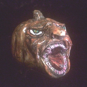 May include: A ceramic pumpkin with a grotesque, toothy grin and green eyes. The pumpkin is painted in shades of brown and has a textured surface.