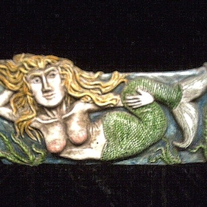 May include: A ceramic wall plaque depicting a mermaid with long blonde hair, a green tail, and a seashell bra. The mermaid is lying on her side with her head resting on her hand. The plaque is decorated with blue and green seaweed.