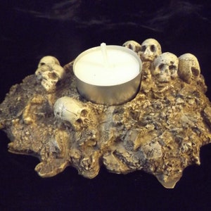 May include: A candle holder with a macabre theme. The holder is shaped like a rough, textured rock formation, adorned with several small, detailed skull sculptures. A single tea light candle sits in the center, providing a soft glow.