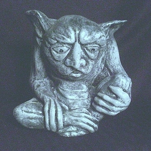 May include: A gray stone sculpture of a gargoyle with a grumpy expression. The gargoyle is sitting with its legs crossed and its hands clasped together.