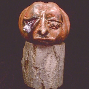 May include: A ceramic pumpkin with a distressed, textured surface. The pumpkin has a human-like face with a missing eye and a large, prominent nose. The pumpkin is sitting on a rough, textured base.