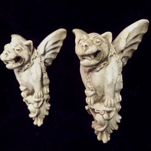 May include: Two white stone gargoyle sculptures with wings, each with a snarling face and detailed carvings. The gargoyles are perched on decorative bases.