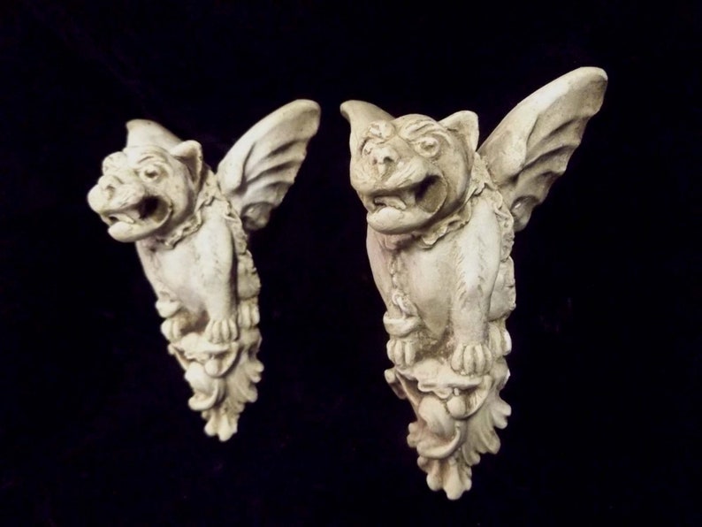 May include: Two white stone gargoyle sculptures with wings, each with a snarling face and detailed carvings. The gargoyles are perched on decorative bases.