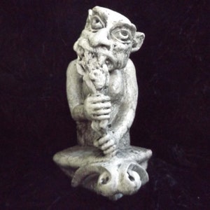May include: A gray stone gargoyle sculpture with a grotesque face and a twisted expression. The gargoyle is holding a branch in its hands and is sitting on a base with a sculpted animal head.