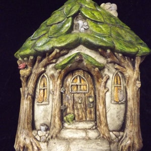 May include: A whimsical fairy house made of sculpted clay with a green leafy roof, brown wooden door, and two windows. The house has a small porch with a frog figurine sitting on the steps.