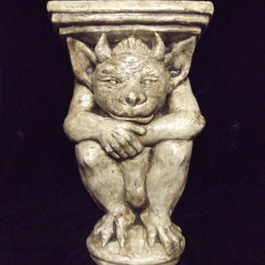 May include: A beige gargoyle sculpture with a shelf on top. The gargoyle is sitting with its hands clasped in front of it. The sculpture is made of a textured material and has a distressed finish.