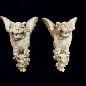 May include: A pair of white stone gargoyle figurines with wings, perched on a decorative base. The gargoyles have a fierce expression with open mouths and sharp teeth.