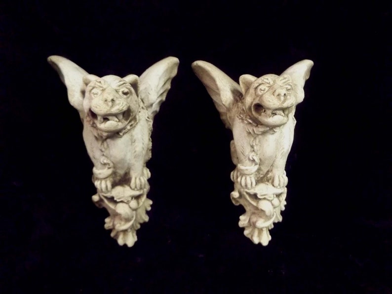 May include: A pair of white stone gargoyle figurines with wings, perched on a decorative base. The gargoyles have a fierce expression with open mouths and sharp teeth.