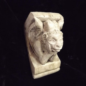 May include: A white stone gargoyle sculpture with wings and a detailed face. The gargoyle is perched on a rectangular base.