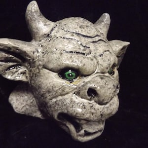 May include: A gray stone gargoyle sculpture with green eyes and a snarling expression. The gargoyle has horns and pointed ears.