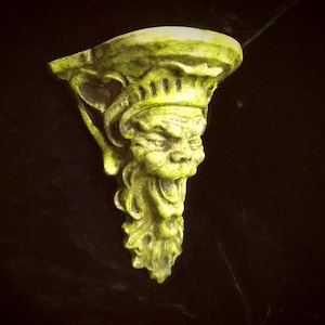 May include: A green, ceramic wall bracket with a grotesque face and a shelf on top. The face has a wide mouth, large eyes, and a pointed nose.