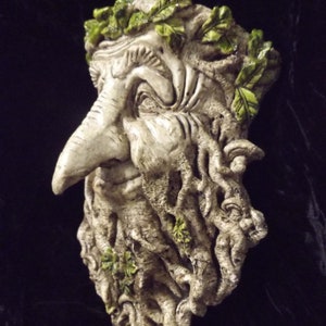 May include: A white, sculpted face with a long nose and a beard made of vines. The face is adorned with green leaves, giving it a woodland or nature-inspired look.