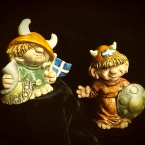 Puede incluir: Dos figuritas de cerámica de gnomos con cascos vikingos. El gnomo de la izquierda sostiene un cuerno y una bandera azul y blanca. El gnomo de la derecha sostiene un escudo con un diseño dorado.