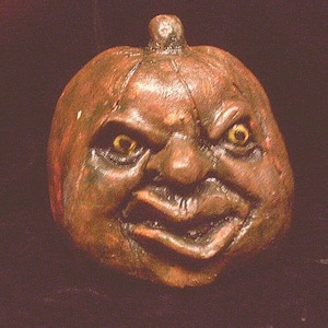 May include: A brown ceramic pumpkin with a carved face, featuring a wide mouth, a large nose, and two yellow eyes. The pumpkin has a textured surface and a dark brown stem.