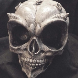 May include: A white alien skull with black eyes and a toothy grin. The skull has small horns on the top and sides.
