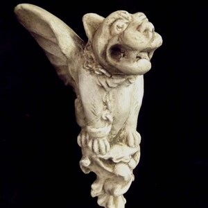 May include: A white stone gargoyle sculpture with wings, a snarling face, and a detailed design. The gargoyle is perched on a stone base.
