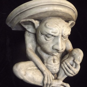 May include: A detailed, off-white gargoyle sculpture with a large, rounded headpiece. The creature has a stern expression, large ears, and is holding a small object. The sculpture is set against a dark background.