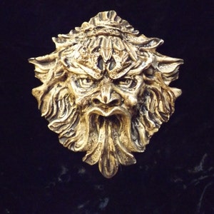 May include: A gold-colored metal sculpture of a grotesque face with a wide open mouth and a long, pointed beard. The sculpture is detailed with intricate carvings and has a textured surface.