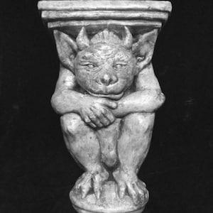 May include: A grayscale statue of a gargoyle with a detailed, textured surface. The gargoyle is seated, with its arms crossed and its legs tucked in. It has horns, large ears, and a stern expression. The statue is mounted on a pedestal.