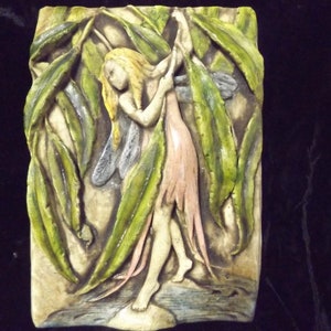 May include: A ceramic wall plaque featuring a fairy sculpted in relief. The fairy is depicted with wings, long flowing hair, and a pink dress. The fairy is surrounded by green leaves.