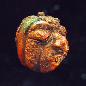 May include: A small, orange pumpkin with a green stem and a grotesque, detailed face. The pumpkin has a cracked and weathered texture, giving it a spooky and eerie appearance.