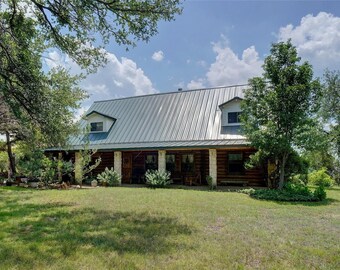 May include: A large, single-story log cabin with a metal roof. The cabin has a porch with wooden columns and a large yard with green grass.