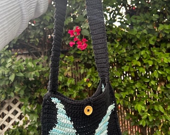 May include: Black crochet shoulder bag with a light blue and green butterfly wing design. The bag has a wooden button closure and a long strap. The bag is held up against a background of green foliage and red flowers.