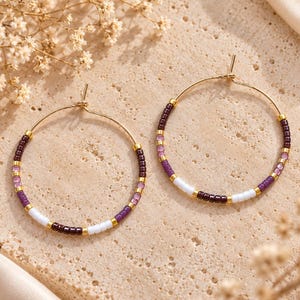 May include: A pair of gold hoop earrings featuring a repeating pattern of small glass seed beads in shades of dark purple, light purple, and white, separated by tiny gold-toned metal spacers along the thin circular wire frame.