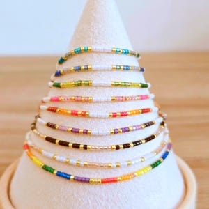 May include: A display of colourful beaded bracelets on a white stand. The bracelets feature small beads in various colours, including blue, green, pink, orange, and gold. Each bracelet has gold accents. The bracelets are delicate and stylish.