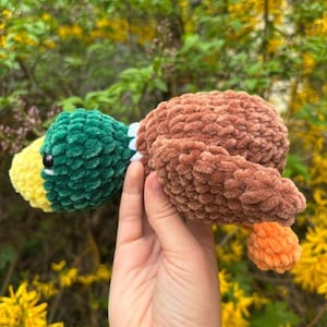 May include: A handmade crochet duck toy. The duck has a green head, yellow beak, and brown body. The duck is made of soft, textured yarn. The toy is held in a hand against a background of green foliage and yellow flowers.