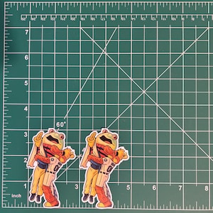 May include: Two stickers featuring a cartoon frog wearing a baseball uniform and giving the middle finger. The frog is yellow, orange, and green. The stickers are on a green cutting mat with inch measurements.