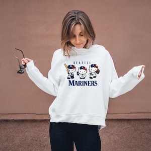 May include: White hoodie featuring a graphic of three cartoon characters in baseball attire with the text "SEATTLE MARINERS". The characters are wearing baseball caps and holding a bat and a baseball glove. The hoodie is a pullover style.