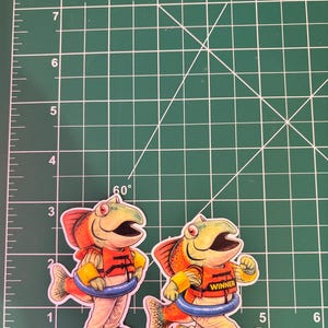 May include: Two stickers of anthropomorphic fish wearing life vests and swim rings. One sticker has the word "WINNER" on the vest. The fish are in a running pose. The background is a green cutting mat with inch measurements.