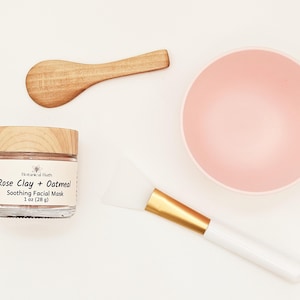 May include: A 1 oz (28 g) glass jar of Botanical Bath Rose Clay + Oatmeal Soothing Facial Mask with a wooden lid, a small wooden scoop, a light pink mixing bowl, and a white silicone facial mask applicator brush with a gold-colored metal ferrule.