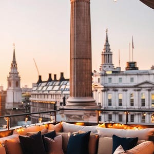May include: A rooftop lounge with a view of the city skyline. The lounge features a large sectional sofa with brown cushions and pillows. The sofa is decorated with fairy lights. The city skyline is visible in the background, including a tall column with a statue on top.